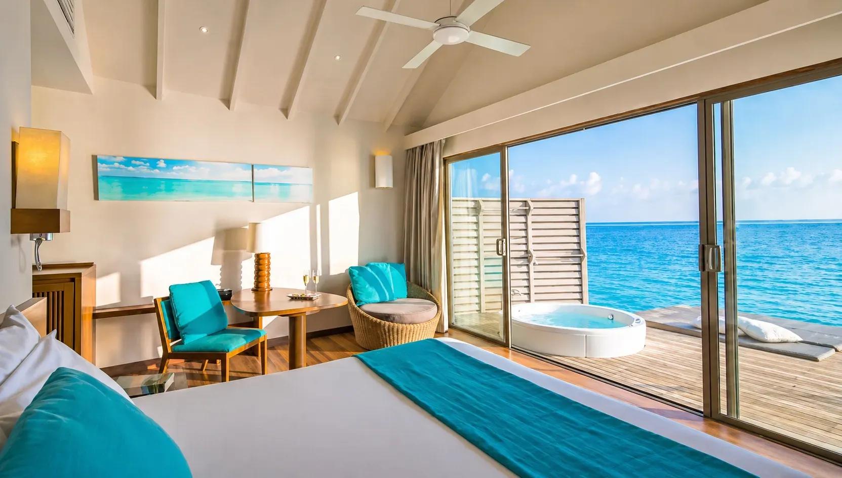 Vista interna della Overwater Villa with Swirl Pool con letto, tavolino e terrazza vista mare al Centara Ras Fushi Resort & Spa Maldives.