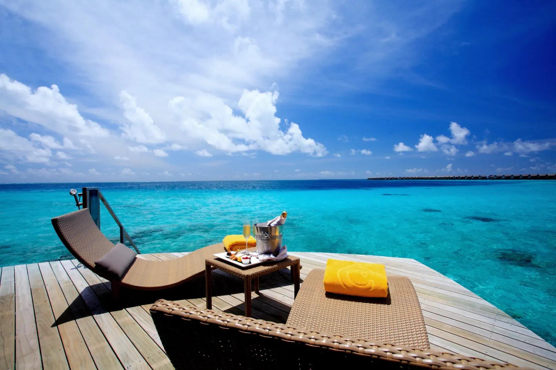 Terrazza della Sunset Overwater Villa con lettini in vimini e vista sull'oceano al Centara Ras Fushi Resort & Spa Maldives.