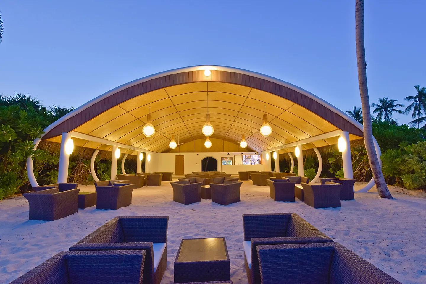 Il Beach Bar del Dreamland The Unique Sea And Lake Resort Spa al crepuscolo, con salottini in vimini disposti sulla sabbia bianca sotto un'ampia tettoia a volta illuminata.