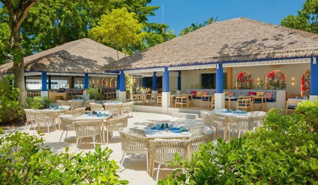 Ristorante all'aperto di Eri Maldives con tavoli apparecchiati sulla sabbia, sedie intrecciate e padiglioni dal tetto in paglia con colonne blu.