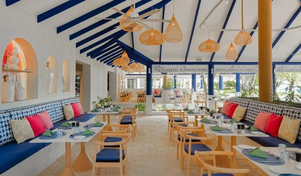 Sala del ristorante di Eri Maldives con pavimento di sabbia, tavoli apparecchiati, divanetti nei toni del blu e soffitto bianco con travi blu.
