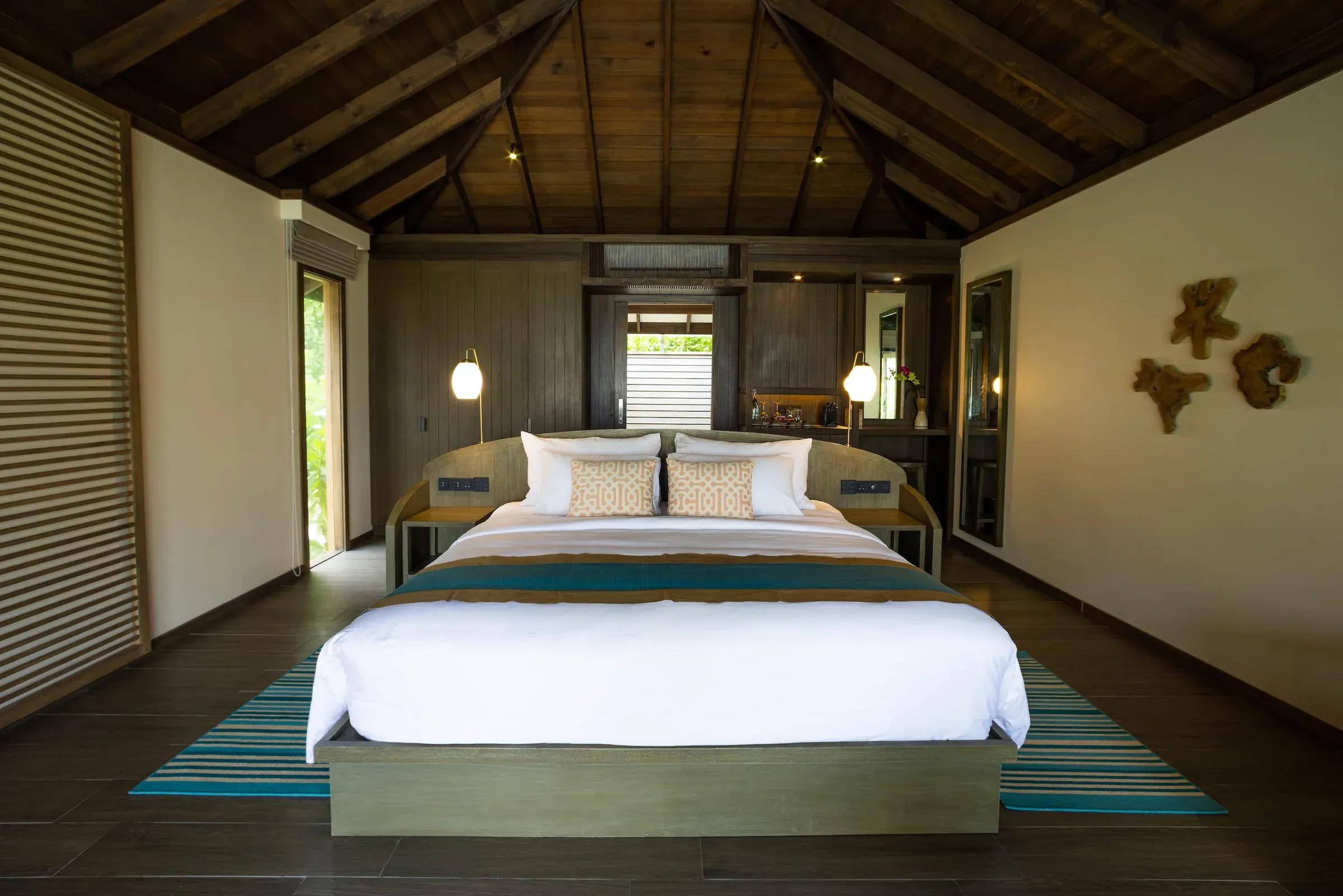 Camera della Dheru Beach Villa con letto matrimoniale, soffitto in legno e illuminazione moderna al Jawakara Islands Maldives.