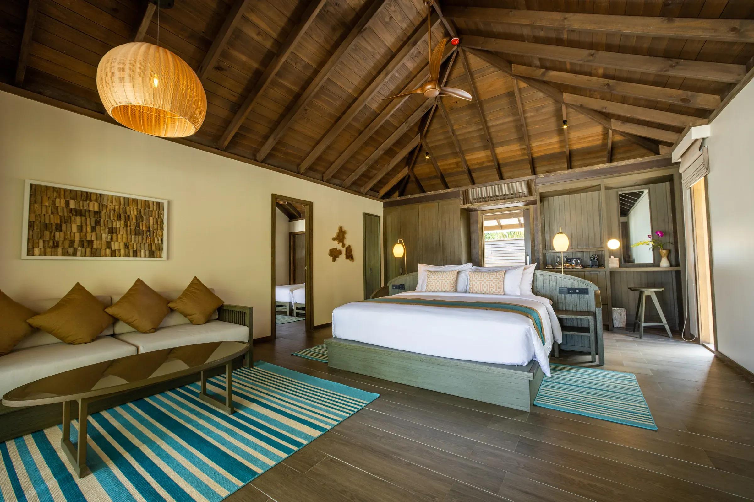 Interno della Dheru Three Bedroom Villa con letto matrimoniale, soffitto in legno e decorazioni calde al Jawakara Islands Maldives.