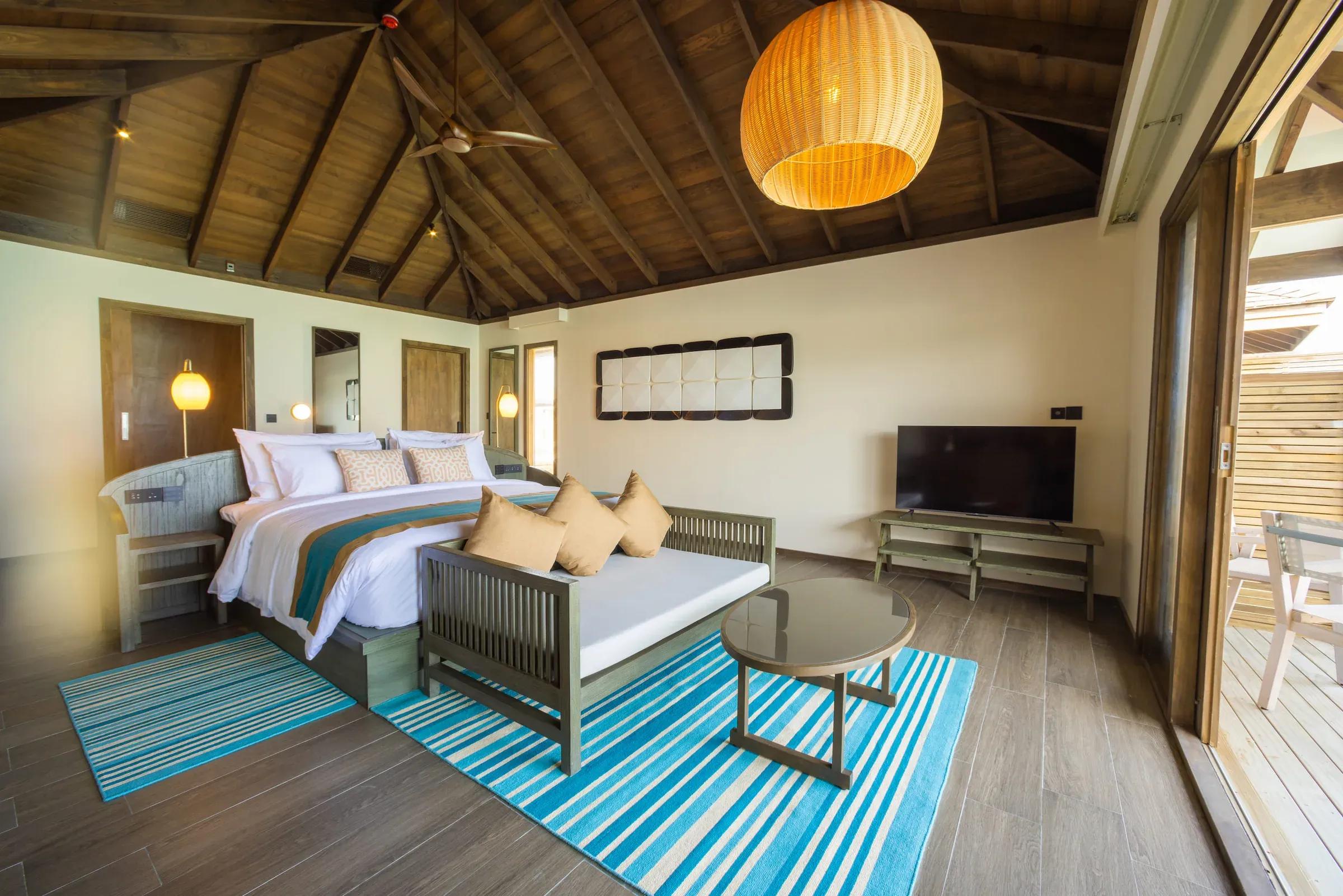 Camera da letto con tetto in legno e area soggiorno della Dheru Water Pool Villa al Jawakara Islands Maldives