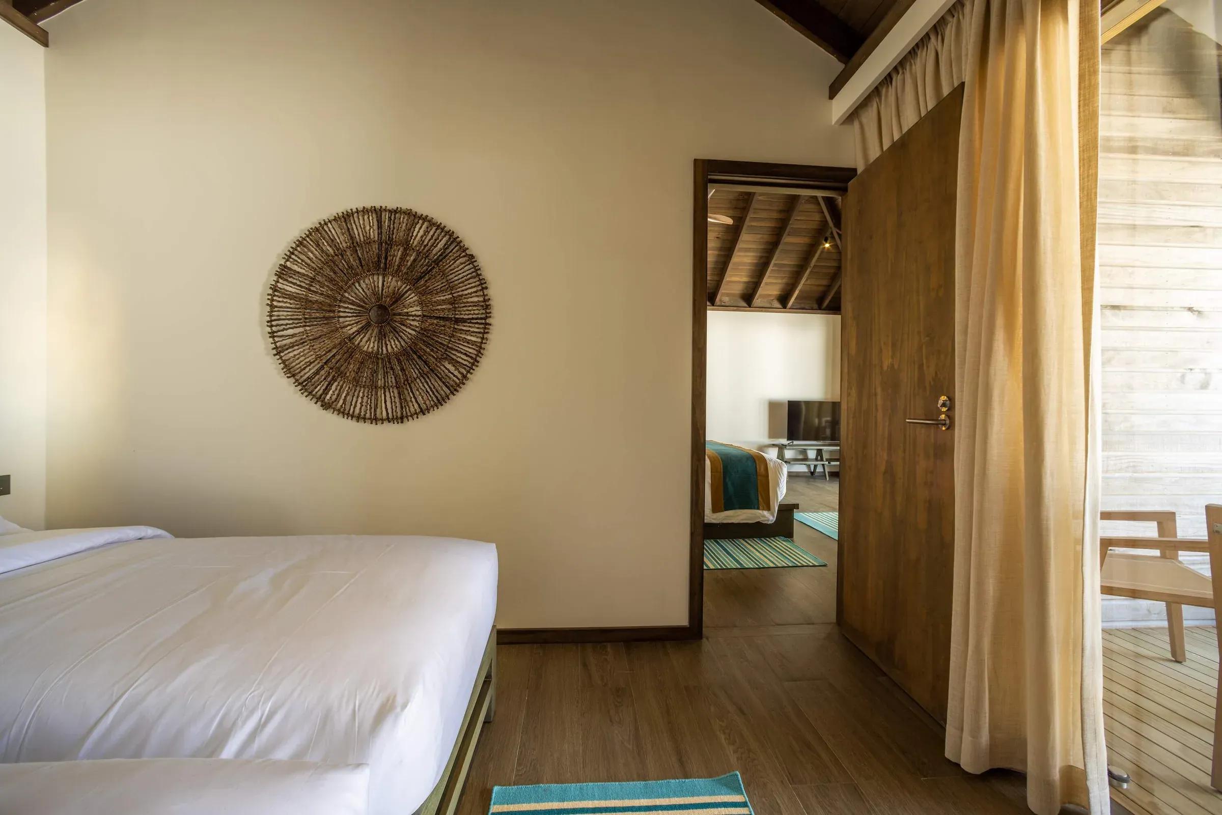 Interno della Mabin Three Bedroom Villa con letto, pareti beige e decorazione in vimini al Jawakara Islands Maldives.
