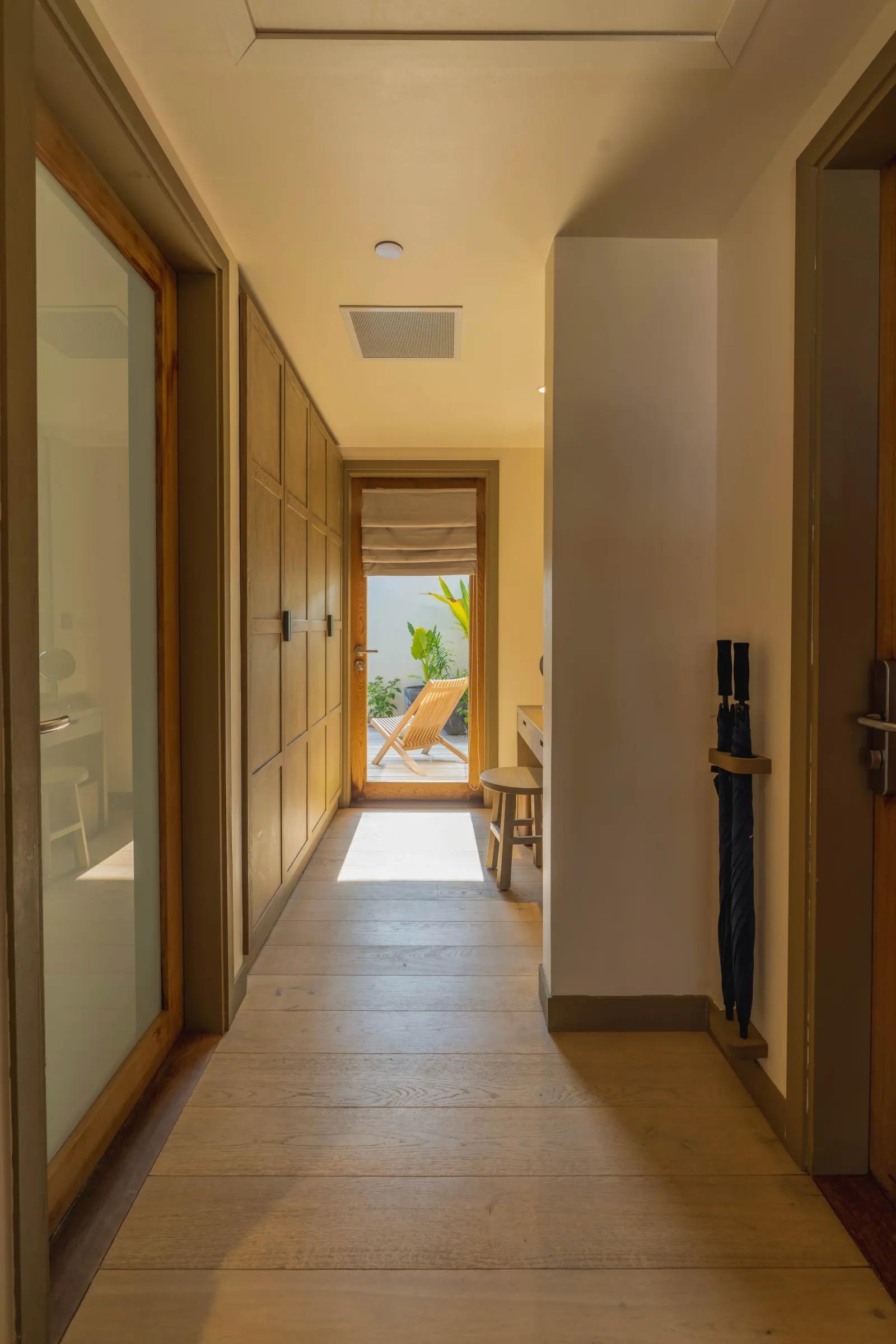 Corridoio d'ingresso della Lagoon Pool Villa al Kagi Maldives Spa Island, con pavimento in legno chiaro, armadi a muro e una porta aperta che conduce a un patio esterno con una sedia a sdraio.