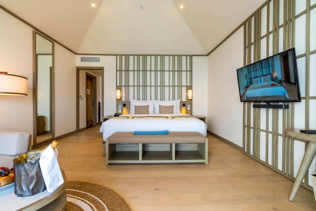 Camera da letto della Overwater Pool Villa al Kagi Maldives Spa Island, con letto king-size centrale, pavimento in parquet, testiera in legno a listelli, TV a schermo piatto e un'area salotto in primo piano.