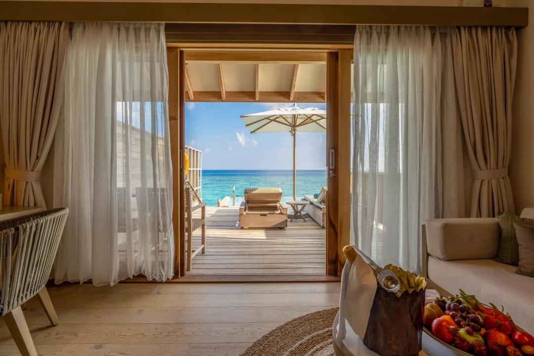 Interno della Overwater Pool Villa al Kagi Maldives Spa Island, con vista dalla porta aperta sulla terrazza privata in legno, attrezzata con lettino e ombrellone, che si affaccia sull'oceano turchese.