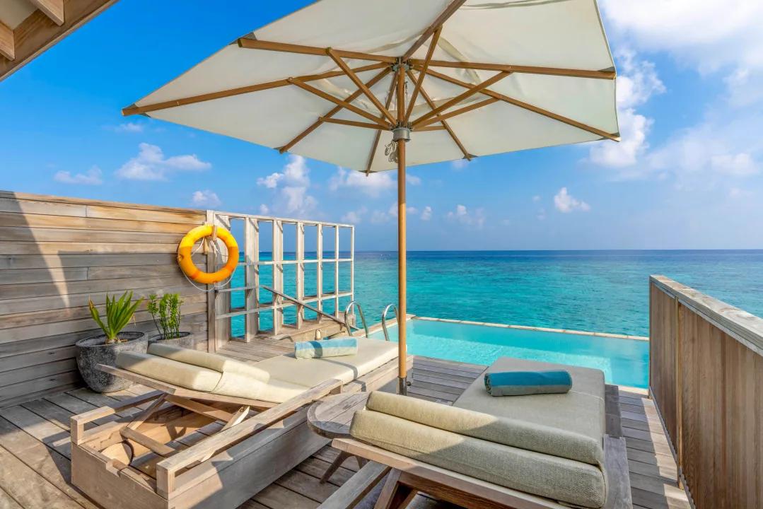 Terrazza esterna in legno della Overwater Pool Villa con piscina privata a sfioro, due lettini prendisole sotto un ombrellone e vista sull'oceano turchese al Kagi Maldives Spa Island.