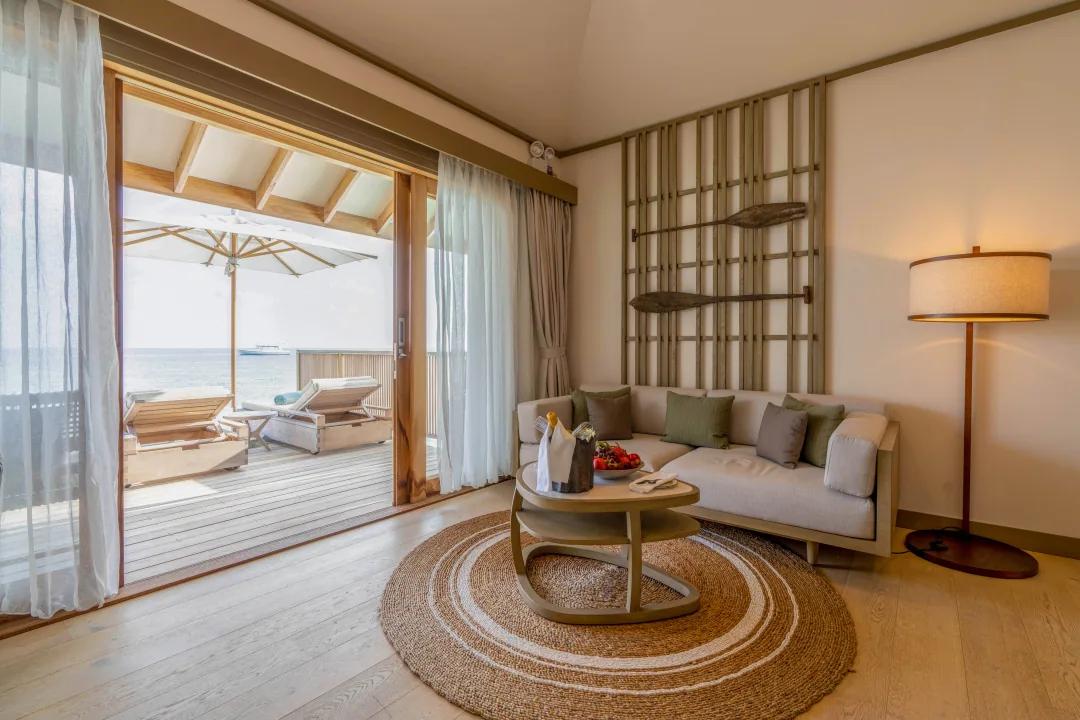 Area soggiorno della Overwater Pool Villa al Kagi Maldives Spa Island, arredata con divano, tappeto circolare, lampada da terra e una porta finestra aperta sulla terrazza esterna con lettini, ombrellone e vista sull'oceano.