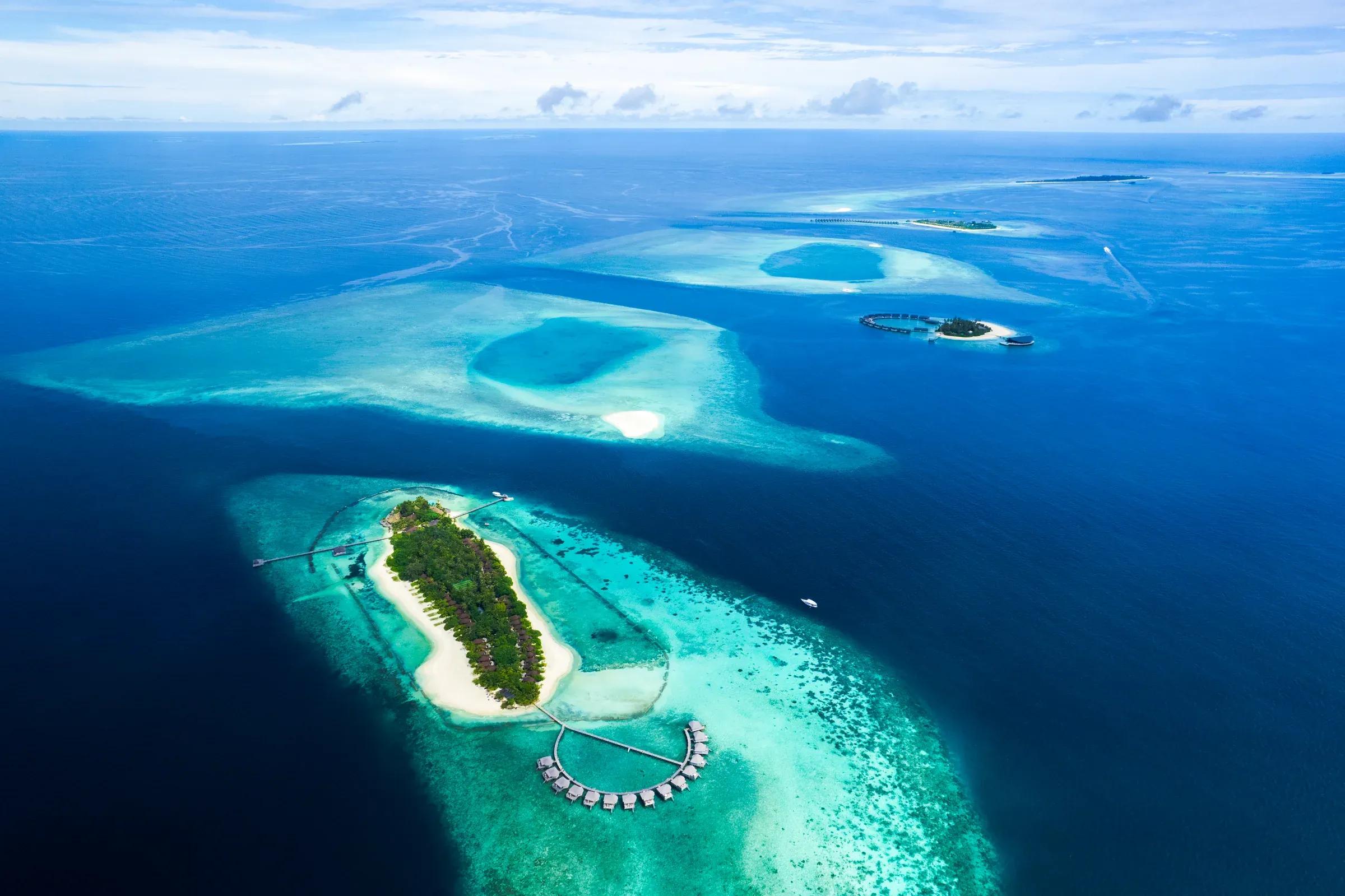 Vista aerea dell'isola tropicale e ville sull'acqua del Komandoo Island Resort & Spa circondate da lagune turchesi.