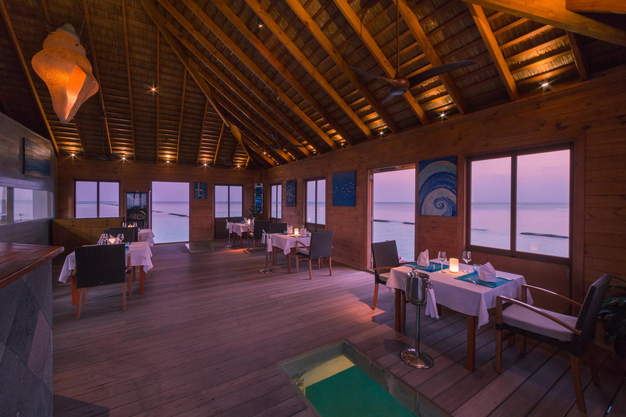 Aqua restaurant con tavoli apparecchiati e vista mare al tramonto al Komandoo Island Resort & Spa.