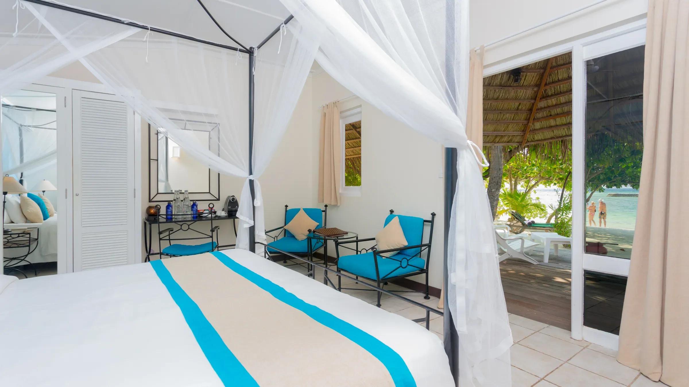 Interno di una Beach Bungalow con letto a baldacchino, sedie blu e vista sulla spiaggia del Kuredu Island Resort & Spa.
