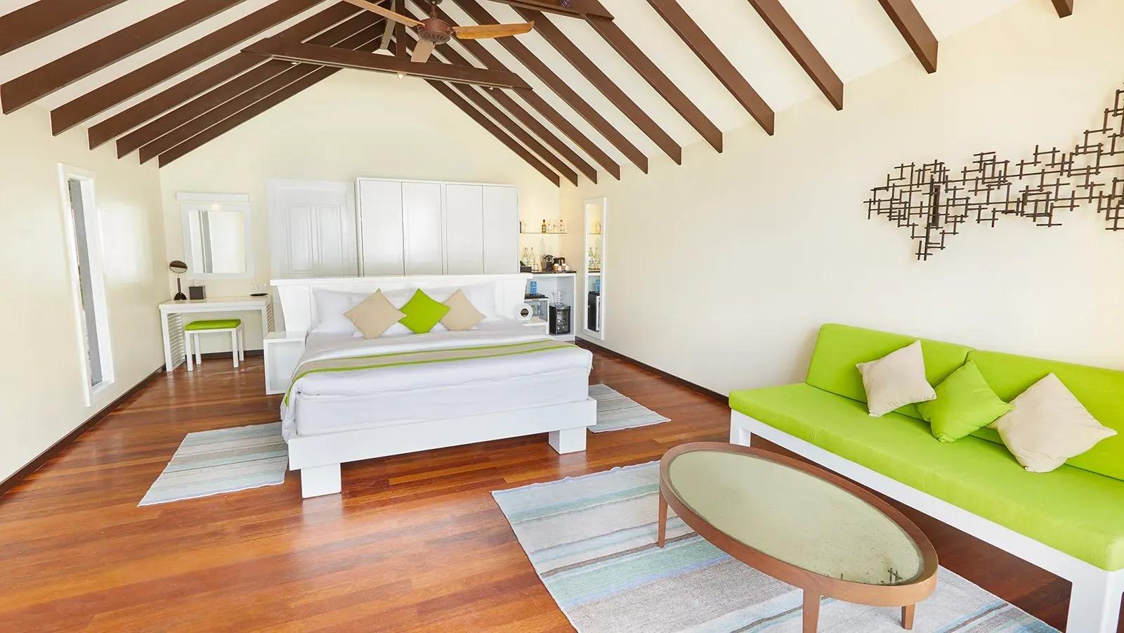 Interno della Deluxe Beach Villa con letto matrimoniale e divano verde al Kuredu Island Resort & Spa.