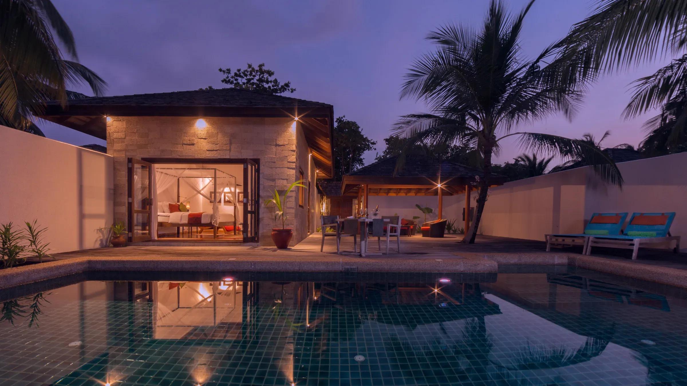 Esterno della Private Pool Villa al Kuredu Island Resort & Spa, ripreso al crepuscolo con la piscina privata illuminata, il patio con lettini, palme e la camera da letto con letto a baldacchino visibile attraverso le porte aperte.