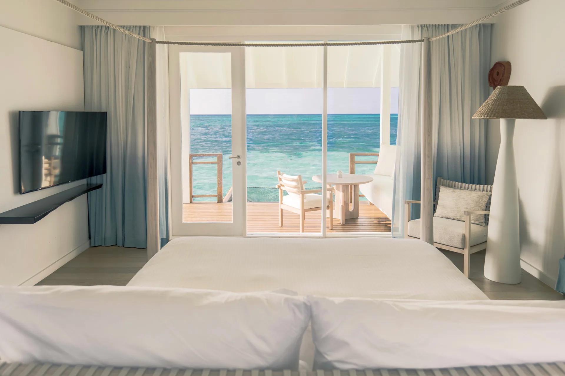 Interno della Water Villa con letto, TV a schermo piatto e vista mare al LUX* South Ari Atoll.
