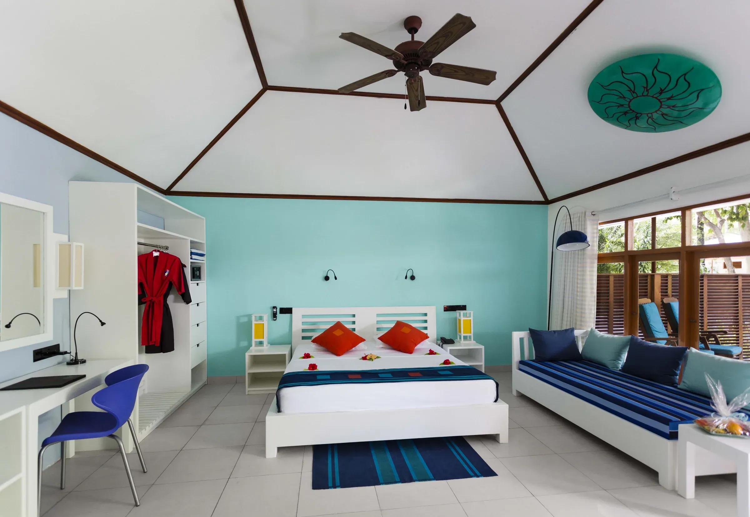 Garden Room con letto matrimoniale e ventilatore a soffitto al Meeru Island Resort & Spa, vista della camera con divano e scrivania.
