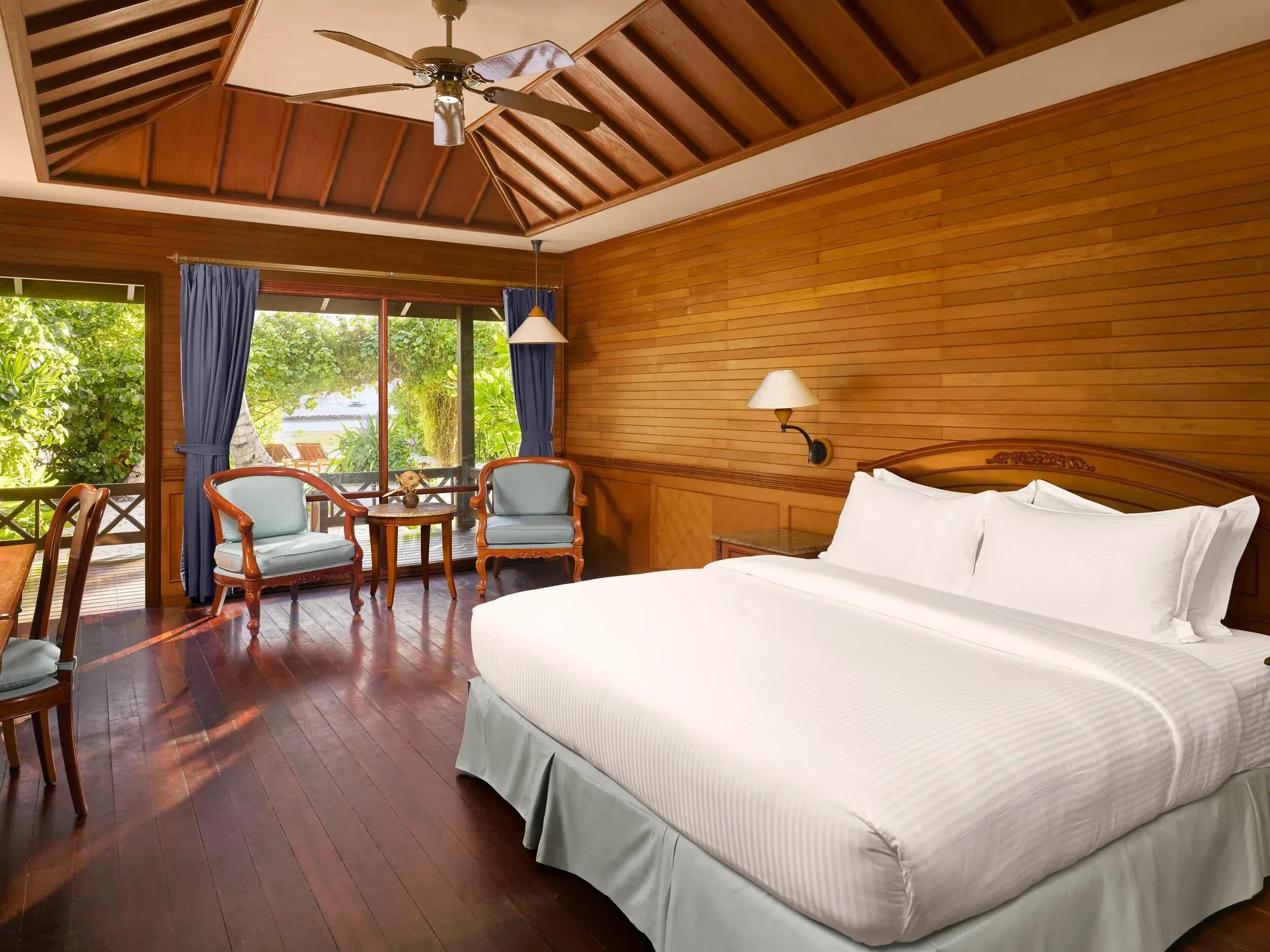 Letto matrimoniale e poltrone nella Sunset Beach Villa del Royal Island Resort & Spa con ventilatore a soffitto e terrazza con vista giardino.