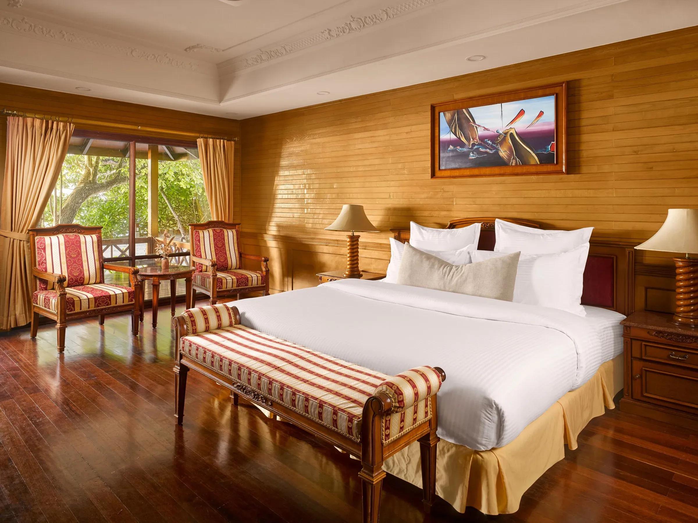 Interno lussuoso della camera da letto nella Two Bedroom Beach Pool Residence, con ampio letto king-size, arredi in legno pregiato, pavimento in parquet e una rilassante vista sul giardino tropicale privato del Royal Island Resort & Spa.