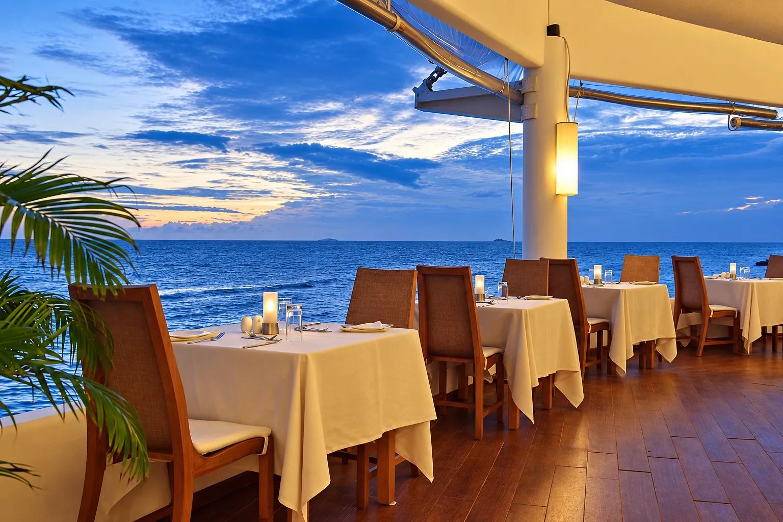 Tavoli apparecchiati per la cena sulla terrazza di un ristorante del Safari Island Resort, affacciata sul mare. La luce del tramonto colora il cielo e l'orizzonte.