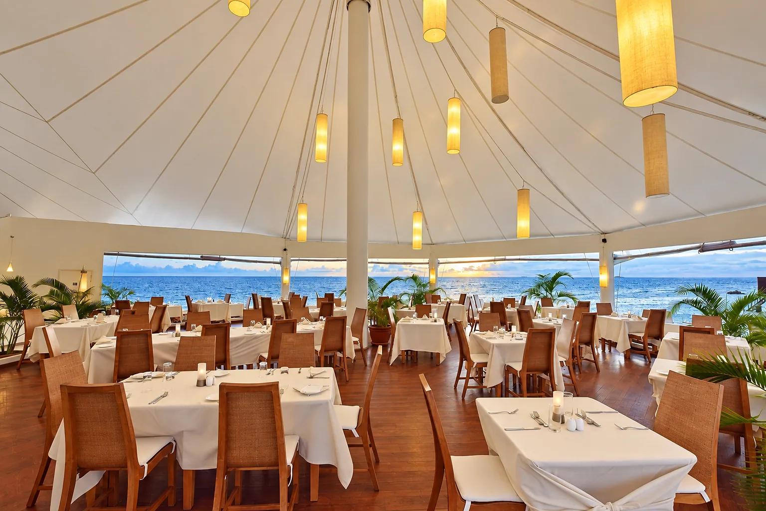 Sala ristorante del Safari Island Resort con soffitto a tenda e vista sul mare. I tavoli con tovaglie bianche sono disposti sul pavimento in legno e illuminati da lampade calde.