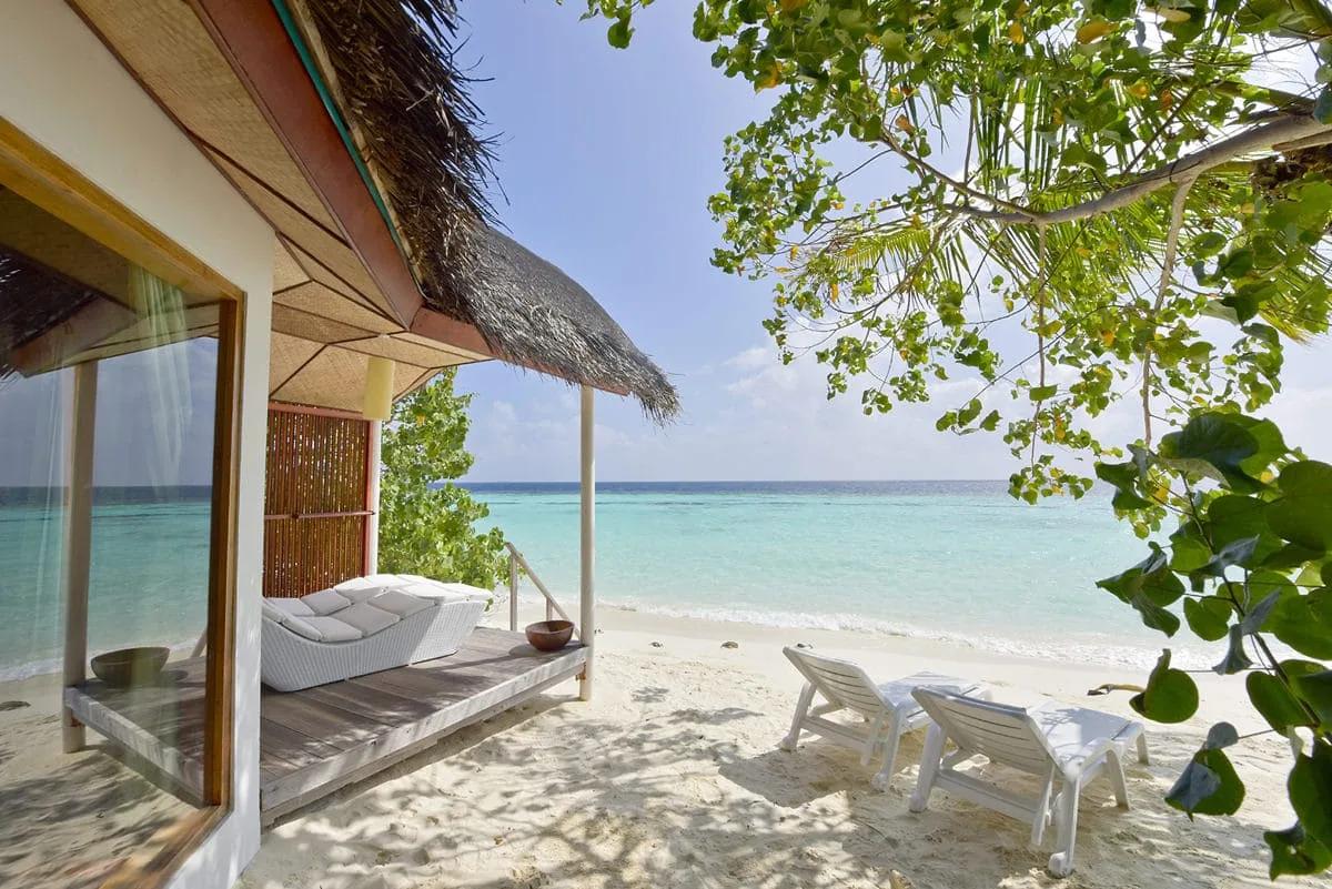 L'esterno di un Beach Bungalow del Safari Island Resort con tetto di paglia, affacciato sulla spiaggia di sabbia bianca. Sulla veranda in legno c'è un lettino e due sdraio sono sulla sabbia, di fronte al mare turchese.