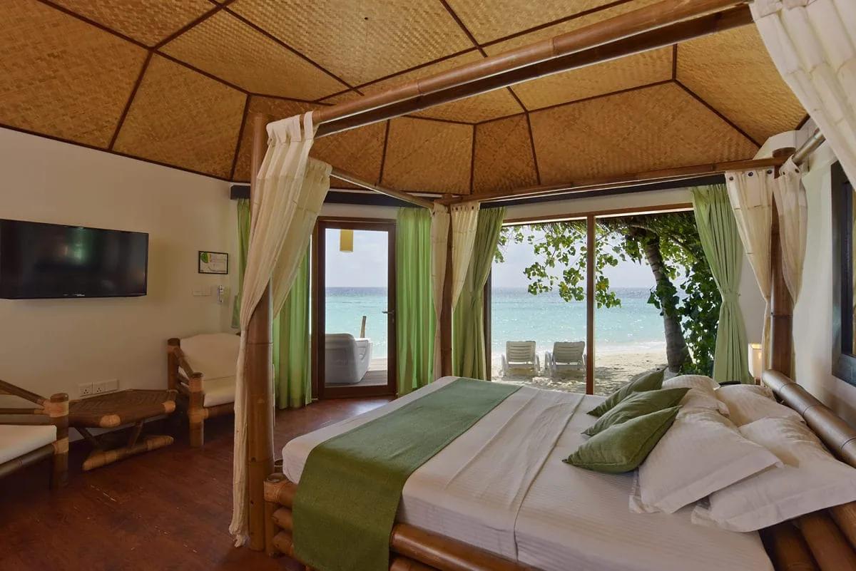 Interno del Beach Bungalow al Safari Island Resort con letto a baldacchino in bambù e soffitto in paglia. La vetrata si apre sulla veranda con vasca e sulla spiaggia con vista mare.