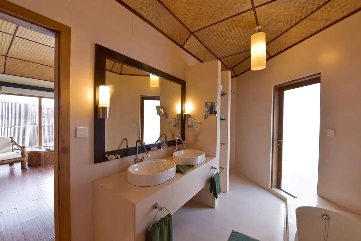 Luminoso bagno del Semi Water Bungalow al Safari Island Resort, con doppio lavabo d'appoggio, specchio con cornice scura e soffitto a volta con pannelli in materiale naturale intrecciato. Si intravede la zona giorno con pavimento in legno.