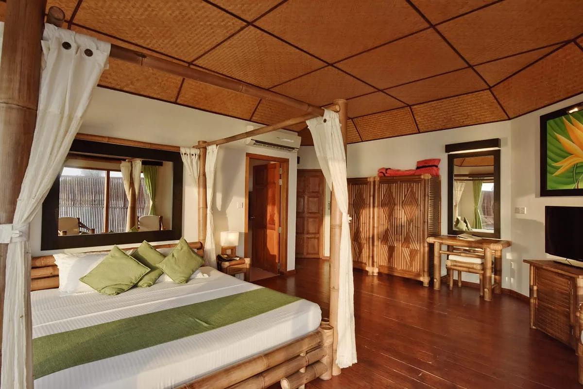 Interno di un Semi Water Bungalow del Safari Island Resort, con un letto matrimoniale a baldacchino in bambù, arredi coordinati, pavimento in legno e soffitto con pannelli intrecciati.