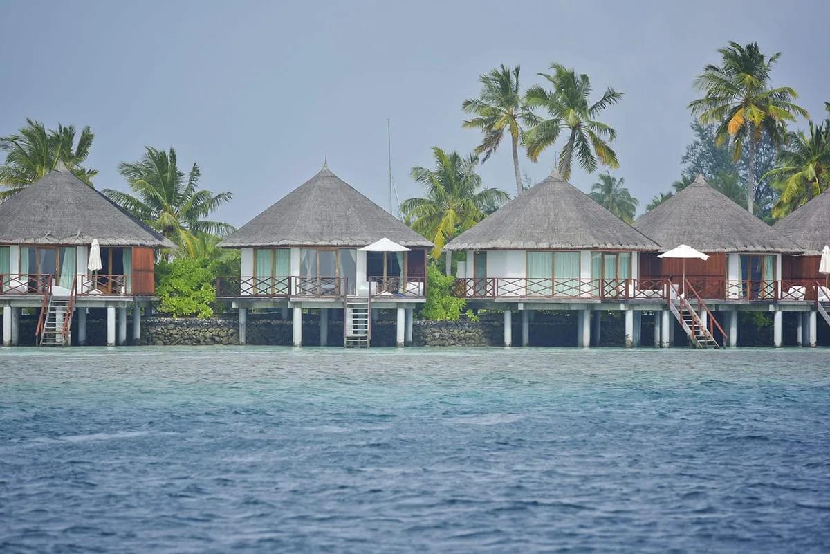 Fila di Semi Water Bungalow del Safari Island Resort visti dal mare, con tetti conici in paglia e terrazze in legno su palafitte. Sullo sfondo, la vegetazione dell'isola con le palme.