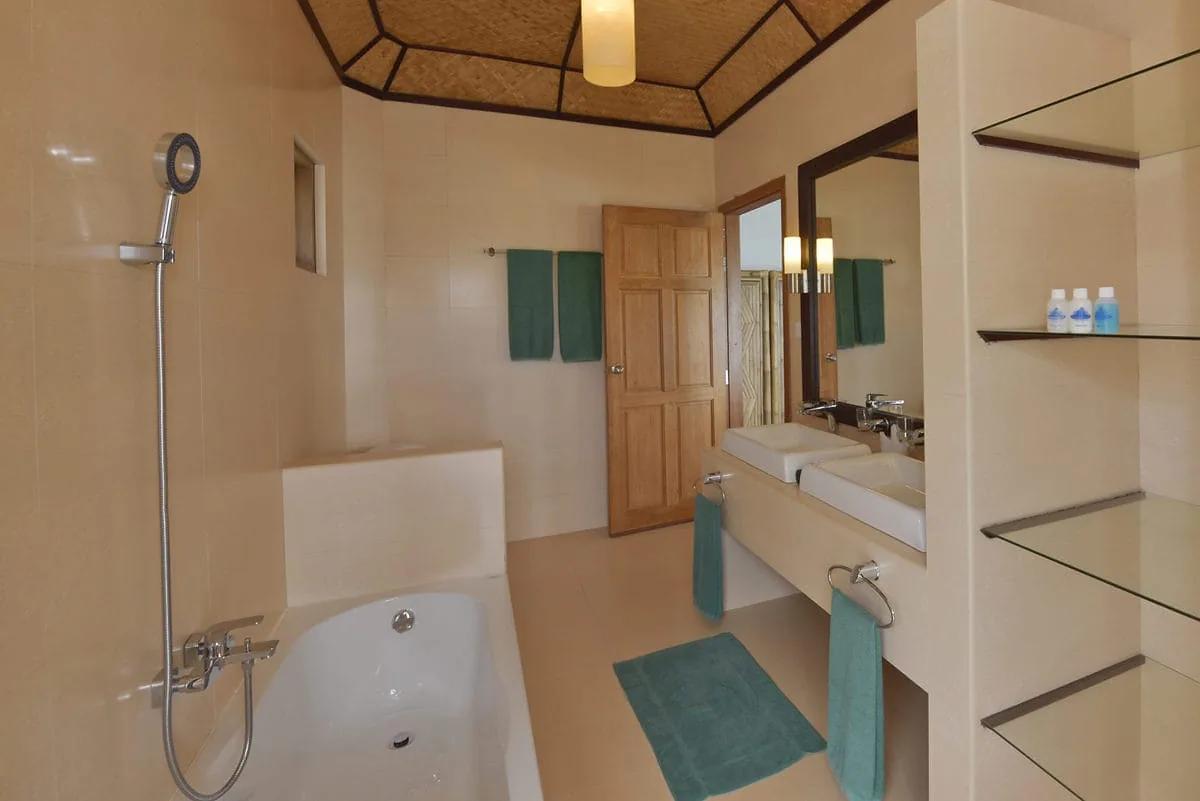 Bagno del Water Bungalow al Safari Island Resort con vasca, doccino, doppio lavabo, porta in legno e soffitto con travi e rivestimento in paglia intrecciata.