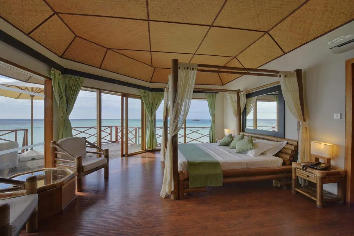 Interno del Water Bungalow del Safari Island Resort con arredi in bambù, letto a baldacchino e soffitto con pannelli intrecciati. Le ampie vetrate si aprono su una terrazza privata affacciata sul mare.