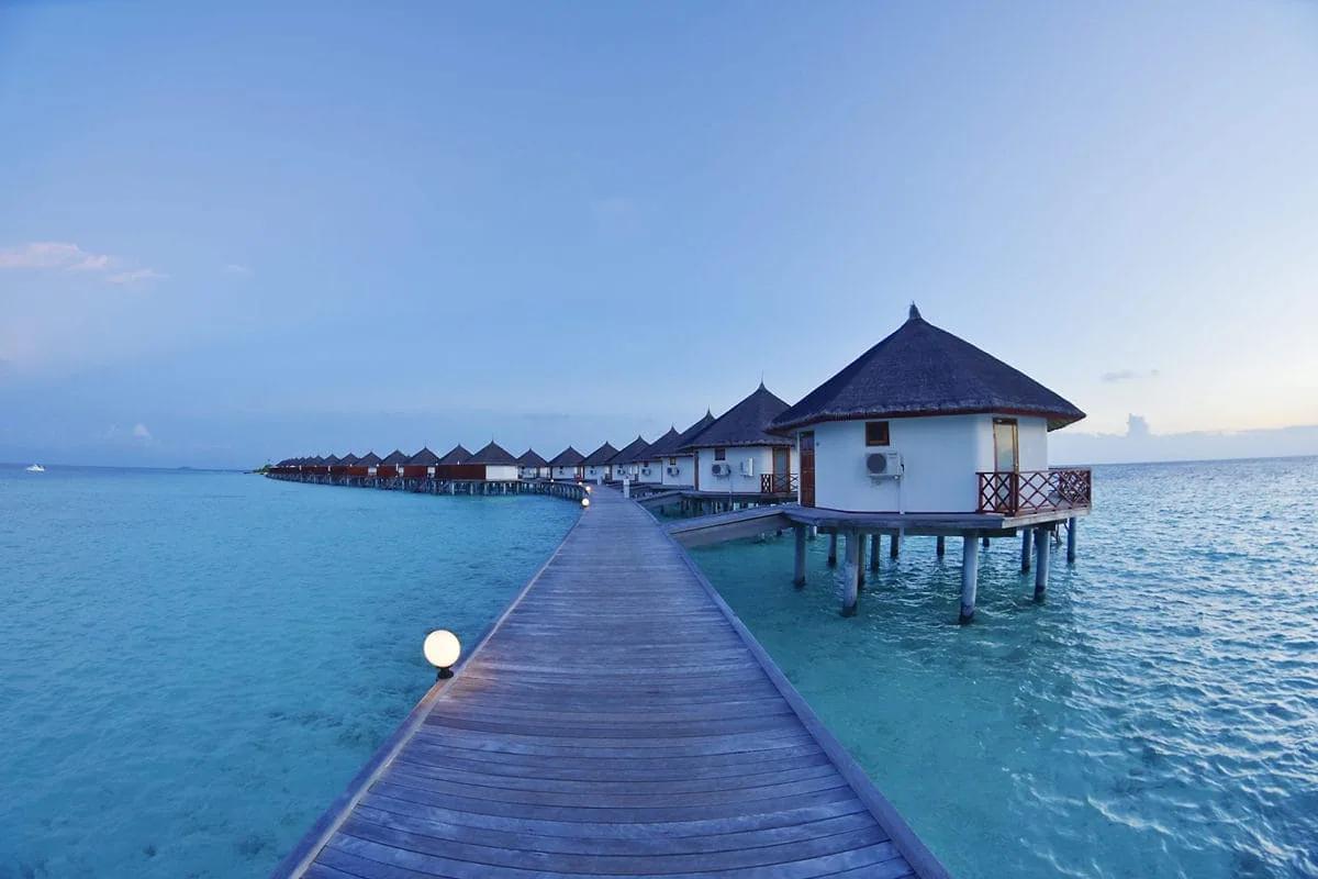 Pontile in legno che si estende sul mare turchese verso la fila dei Water Bungalow del Safari Island Resort, palafitte con tetti di paglia illuminate dalla luce del crepuscolo.