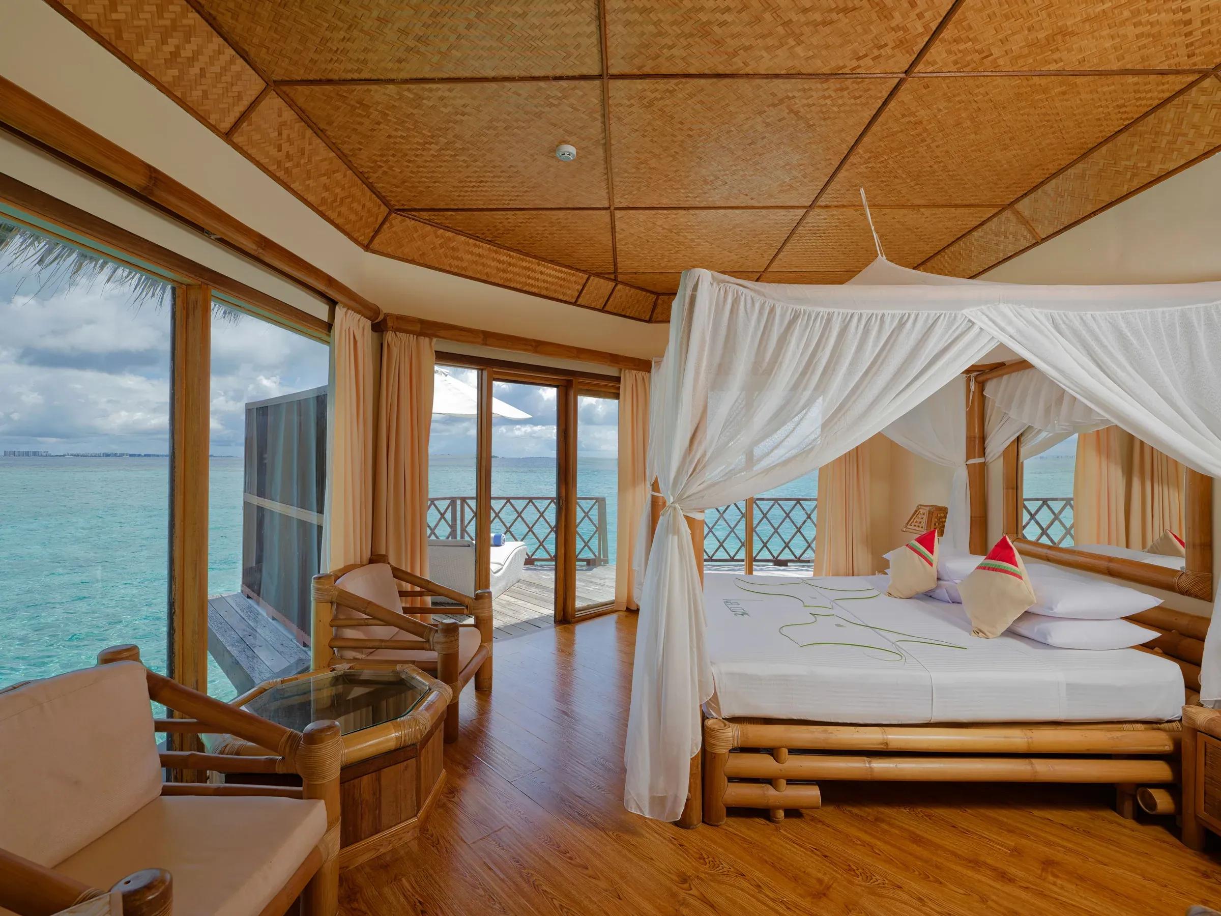 Interno della Water Bungalow con letto a baldacchino, sedie in legno e vista sull'oceano al Thulhagiri Island Resort & Spa.