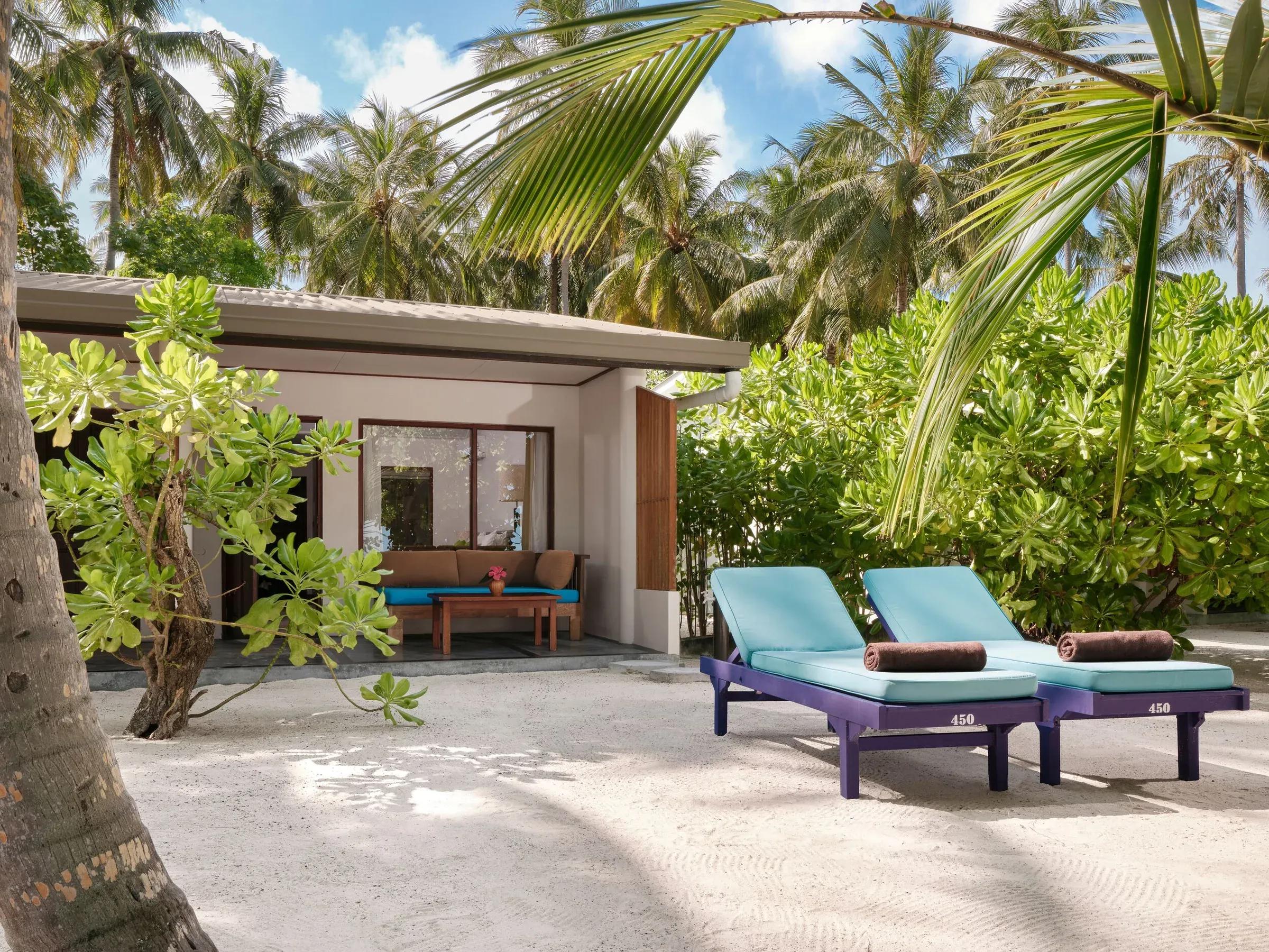 Sunset Beach Villa al Villa Park Sun Island Resort & Spa, con eleganti lettini blu sulla sabbia bianca, circondati da rigogliose palme tropicali e vegetazione lussureggiante.