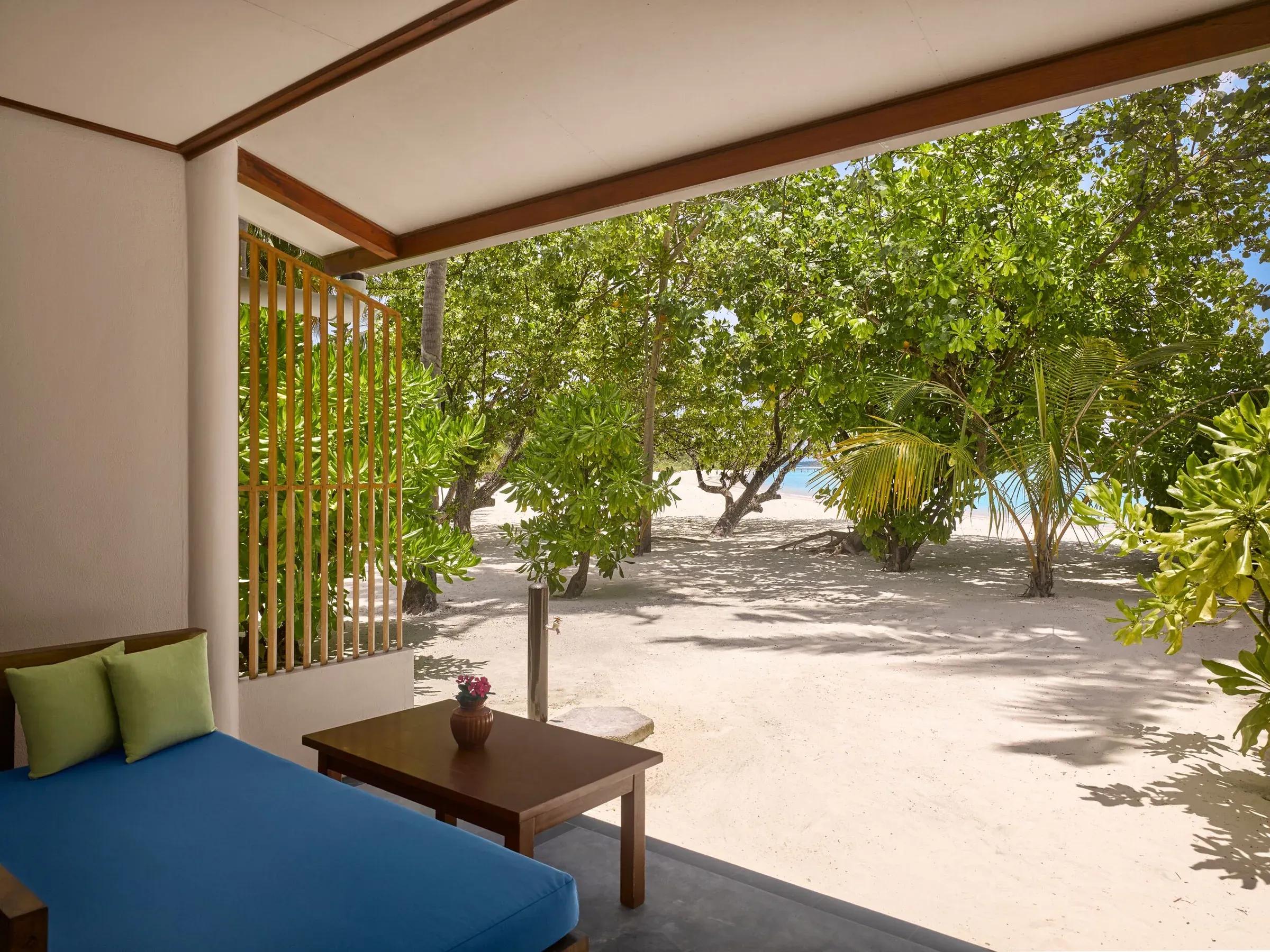 Vista dalla Sunset Beach Villa del Villa Park Sun Island Resort & Spa, con divano all'aperto su una terrazza che dà su una spiaggia dorata e alberi rigogliosi, con l'oceano appena visibile oltre il fogliame.